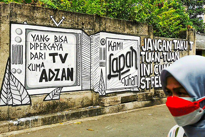 Warga saat melintas dengan berlatar belakang mural yang bertuliskan 'kami lapar tuhan' di Jakarta Pusat, Selasa (24/8/2021).